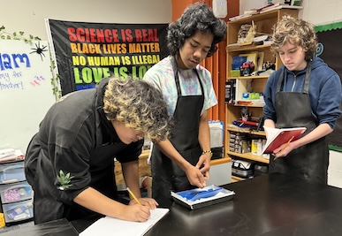 3 IVA High Students work on a science lab. One student is writing in a notebook, one is reading out of a notebook and another is listening and getting supplies ready